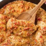 A wooden spoon lifts a piece of creamy Roasted Red Pepper Chicken breast topped with diced tomatoes and herbs from a skillet filled with similar pieces in a rich, seasoned sauce.