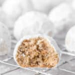 A close-up of a Snowball Cookie, coated in powdered sugar with a bite taken out, reveals its nutty, crumbly interior. More Snowball Cookies are blurred in the background on a cooling rack.