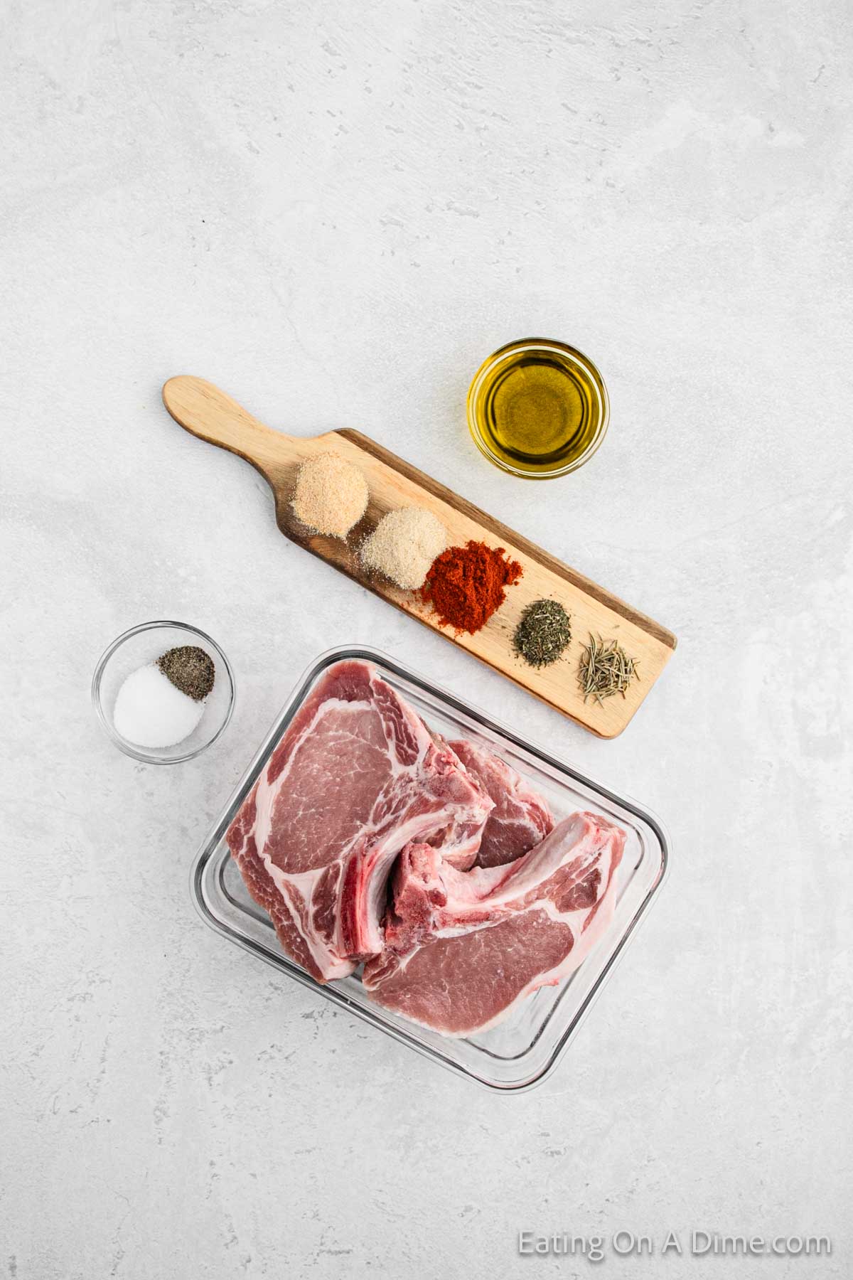 Top-down view of raw bone-in pork chops in a glass dish, placed next to a small wooden board holding five different spices. A small bowl of salt, a small bowl of olive oil, and text at the bottom right corner that says "Eating On A Dime.com" are also visible.