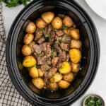 A Crockpot Steak and Potatoes dish, filled with tender beef chunks and whole baby potatoes, is garnished with chopped herbs. It sits on a white surface next to a striped towel and a small bowl of fresh parsley.