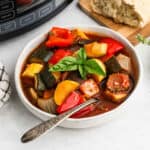 A bowl of colorful Slow Cooker Ratatouille with zucchini, bell peppers, tomatoes, and onions, garnished with fresh basil, with a spoon in the bowl and bread on a wooden board in the background.