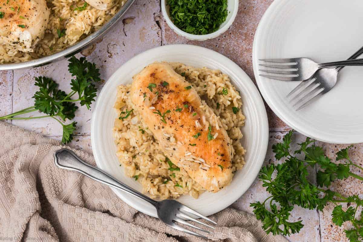 A white plate with creamy rice topped with a seasoned chicken breast, garnished with chopped parsley. Surrounding the dish are sprigs of fresh parsley, a small bowl of chopped parsley, a linen cloth, and an empty white plate with two forks on it.