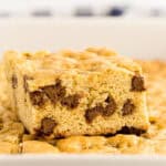 A close-up of a thick, golden-brown blondie bar filled with chocolate chips, resting on top of a pan of similar bars. The texture looks soft and chewy—just like your favorite Chocolate Chip Cookie Cake Recipe in bar form.
