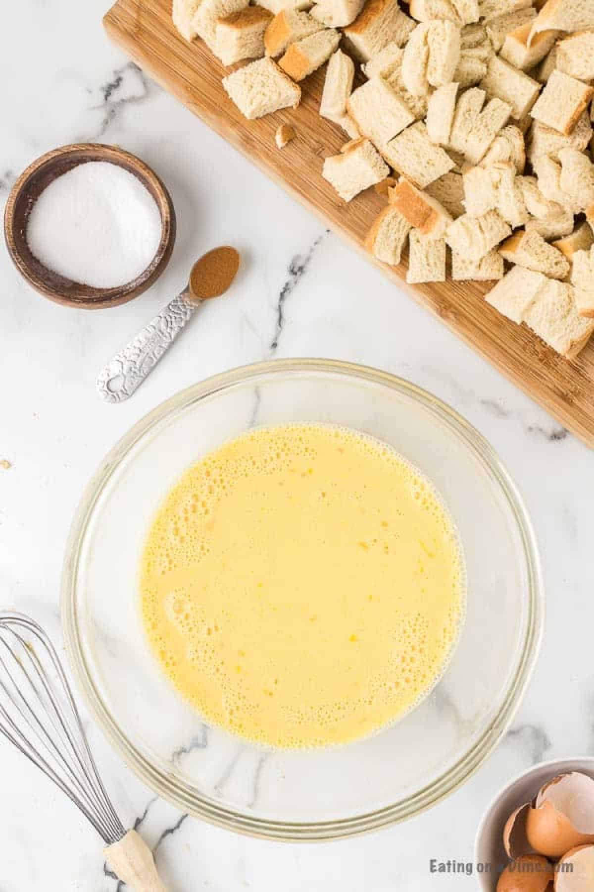 Egg mixture in a bowl with a small bowl of sugar and a teaspoon of cinnamon and a cutting board with pieces of bread