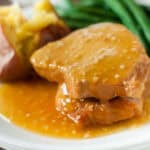 Two pieces of pork chops, inspired by our crock pot honey garlic pork chops recipe, are covered in golden brown gravy on a white plate, with a baked potato and green beans in the background.
