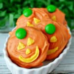 Three Jack-O-Lantern Chocolate Covered Oreos, decorated with orange icing, yellow icing for facial features, and a green candy on top, are arranged in a white dish.