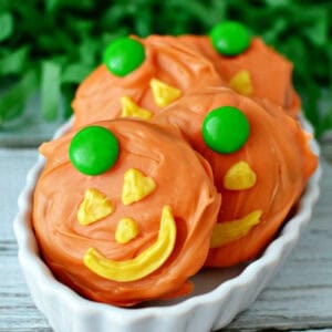 Three Jack-O-Lantern Chocolate Covered Oreos, decorated with orange icing, yellow icing for facial features, and a green candy on top, are arranged in a white dish.