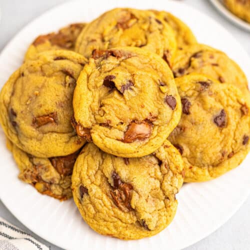 A plate of golden brown Milky Way Cookies piled on top of each other, with visible chunks of melted chocolate throughout.