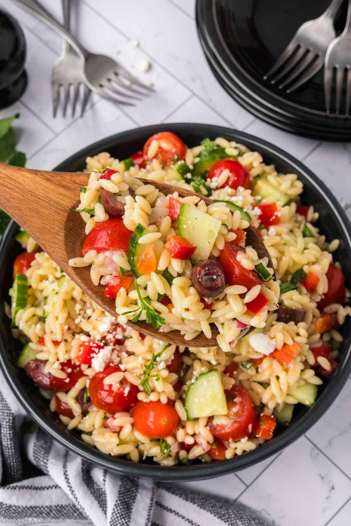 A wooden spoon lifts a serving of vibrant Orzo Pasta Salad with cherry tomatoes, cucumbers, olives, red peppers, and fresh herbs from a black bowl. Plates and forks are visible in the background.