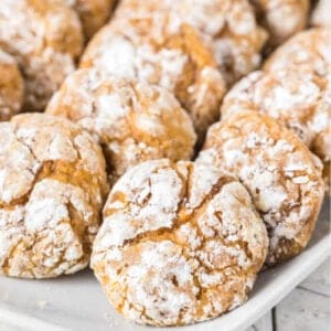 A close-up of several crinkle cookies covered in powdered sugar, arranged on a white plate. These easy pumpkin cookie recipe treats feature a cracked surface and golden brown color.