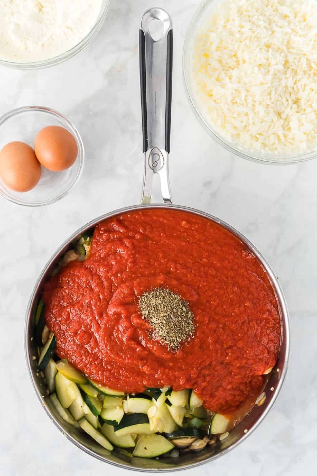 Chopped zucchini and squash in a saucepan topped with marinara sauce with a side of eggs, shredded cheese in bowls