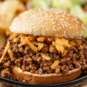 A close-up of a BBQ sloppy joes recipe featuring seasoned ground beef and shredded cheddar cheese piled high on a sesame seed bun.