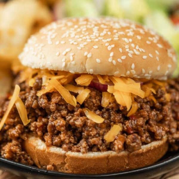 A close-up of a BBQ sloppy joes recipe featuring seasoned ground beef and shredded cheddar cheese piled high on a sesame seed bun.