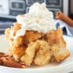 A serving of crock pot bread pudding recipe topped with whipped cream sits on a white plate, with pecan halves beside it and a blurred kitchen appliance in the background.