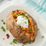 A Crock Pot Baked Potato topped with shredded cheddar cheese, crispy bacon bits, chopped green onions, and a dollop of sour cream sits on a white plate. Extra green onions and bacon pieces are scattered around for garnish.