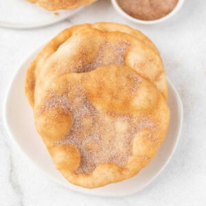 Two pieces of golden fried dough from our Elephant Ears Recipe are dusted with cinnamon sugar and stacked on a white plate, with a small bowl of extra cinnamon sugar in the background.