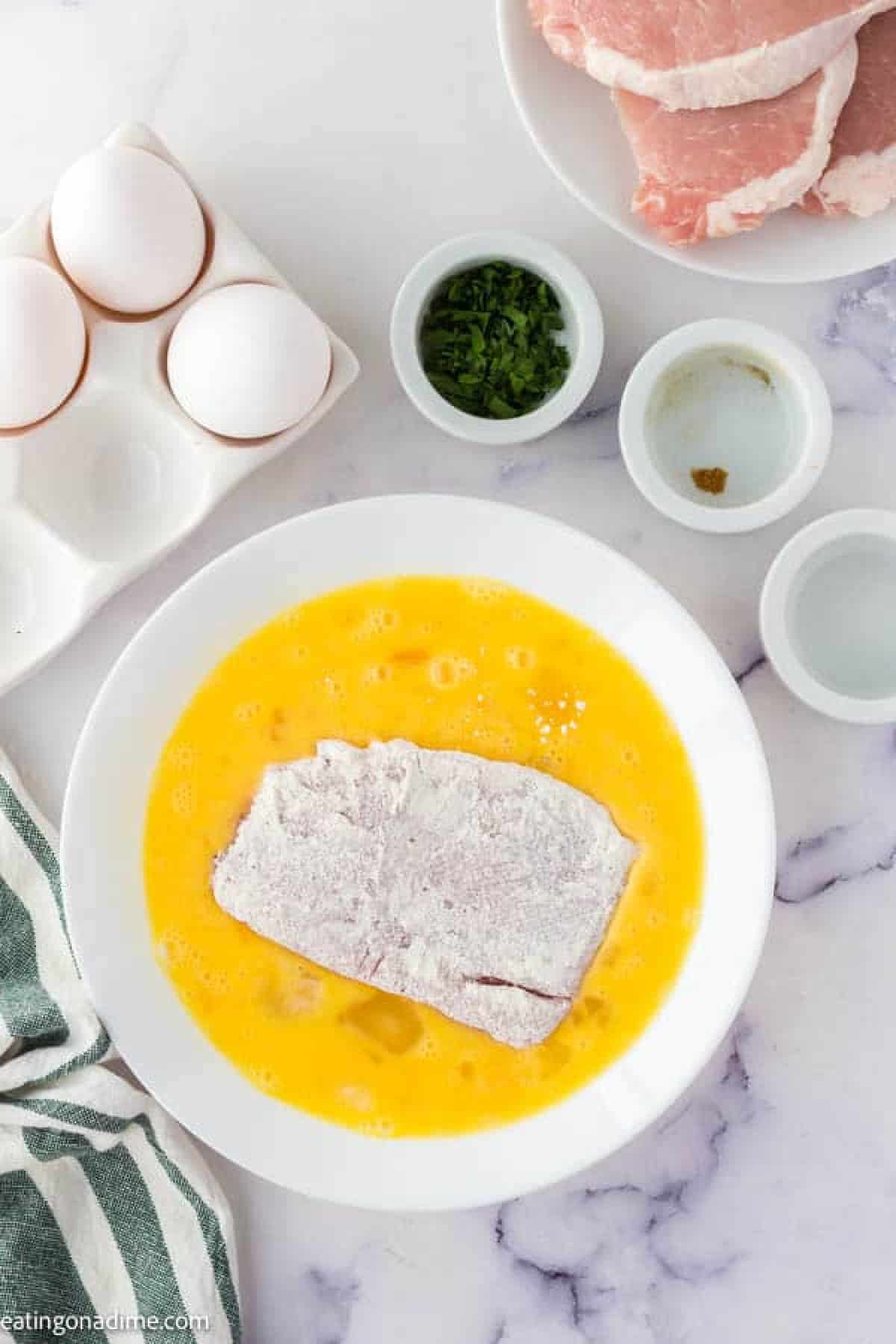 Dipping the floured pork chops in whisked eggs in a bowl with a side of eggs and chopped parsley