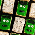 Halloween Rice Krispie Treats shaped like rectangles are decorated as green Frankenstein faces with candy eyes and black icing mouths, and white mummy faces with icing bandages and candy eyes, all arranged on a dark surface.