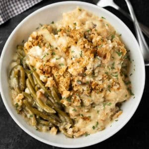 A bowl of creamy Instant Pot Chicken and Stuffing Casserole topped with golden breadcrumbs, served with cooked green beans and garnished with chopped parsley. A fork and spoon are placed nearby.