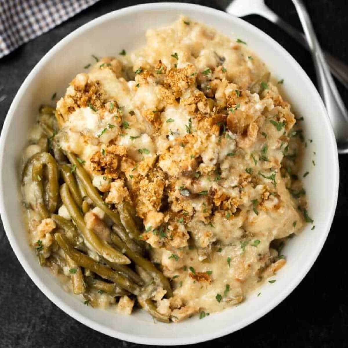 A bowl of creamy Instant Pot Chicken and Stuffing Casserole topped with golden breadcrumbs, served with cooked green beans and garnished with chopped parsley. A fork and spoon are placed nearby.