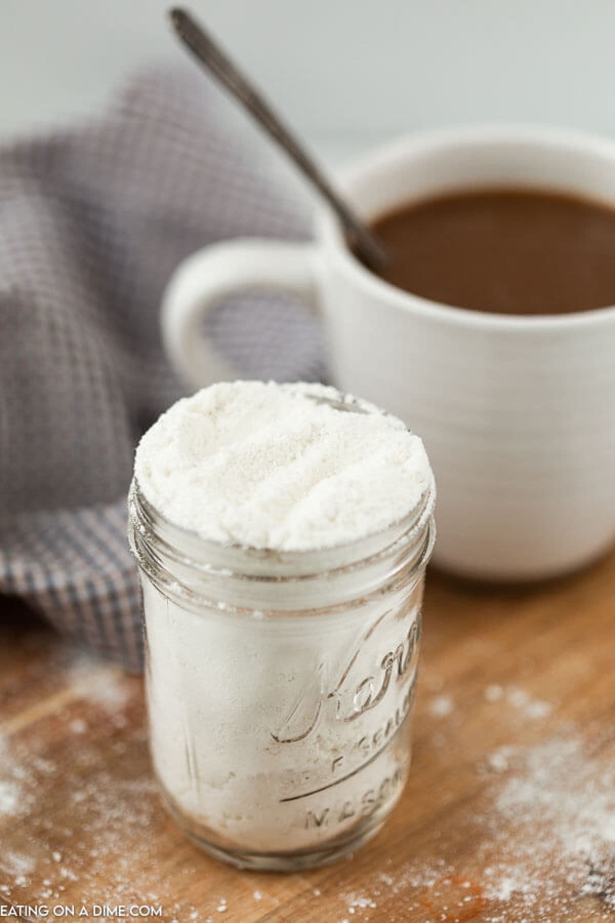 Powdered coffee cream in a jar with a cup of coffee with a spoon in the background