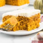 A close-up of a slice of pumpkin cake with white frosting on a white plate. Reminiscent of Paula Deen Pumpkin Bars, a fork has taken a bite from the moist, orange cake. The background shows more cake and part of a striped cloth.
