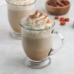 A clear glass mug filled with a creamy Starbucks Pumpkin Spice Latte recipe drink, topped with whipped cream and a sprinkle of cinnamon, sits on a light surface. Another mug and a small wooden bowl with dried berries are in the background.