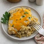 A plate with baked Chicken and Rice Casserole, topped with melted cheddar cheese and served over seasoned rice. A sprig of parsley garnishes the side, while a silver fork rests nearby and salt and pepper shakers sit in the background.
