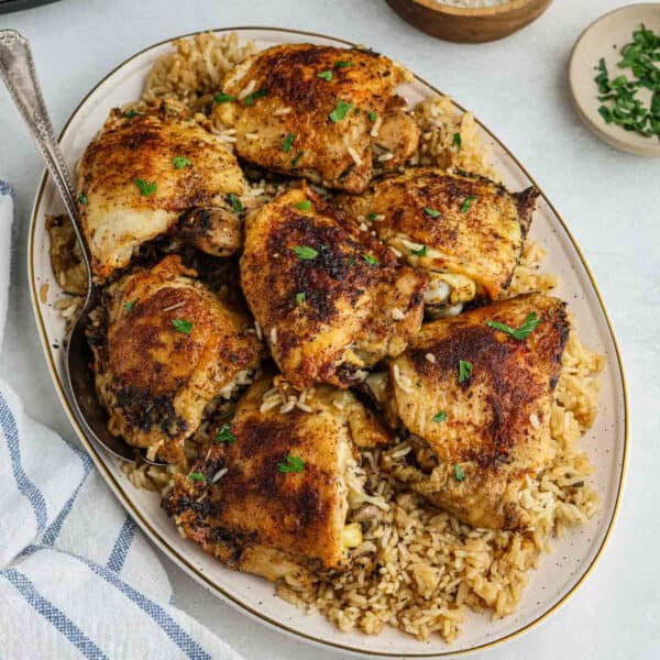 A platter of crispy, seasoned Crock Pot Chicken Thighs and Rice is served over a bed of rice, garnished with chopped parsley. A serving spoon and a cloth napkin are nearby.