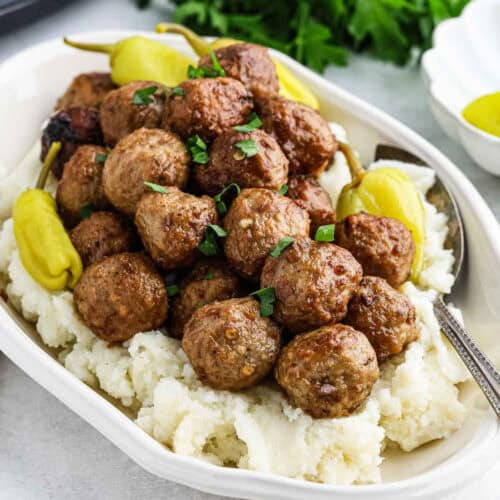 A platter of Crock Pot Mississippi Meatballs garnished with chopped parsley, served over creamy mashed potatoes and accompanied by whole pepperoncini peppers. A serving spoon rests on the side of the dish.