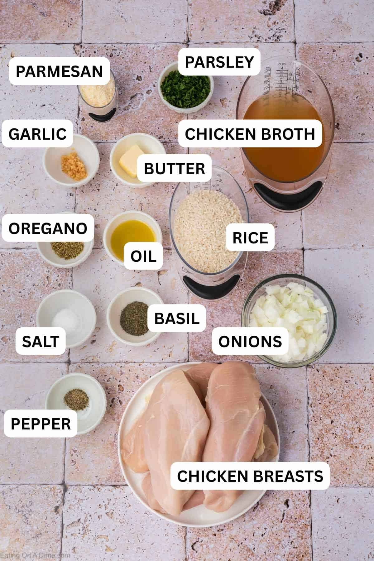 An assortment of ingredients on a tiled surface including raw chicken breasts on a plate, rice, chopped onions, olive oil, butter, grated cheese, minced garlic, chicken broth, and various seasonings in small bowls.