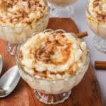 A glass bowl filled with creamy rice pudding, topped with a swirl of cinnamon, sits on a wooden board with a spoon and cinnamon stick nearby. Other bowls of rice pudding can be seen in the background.