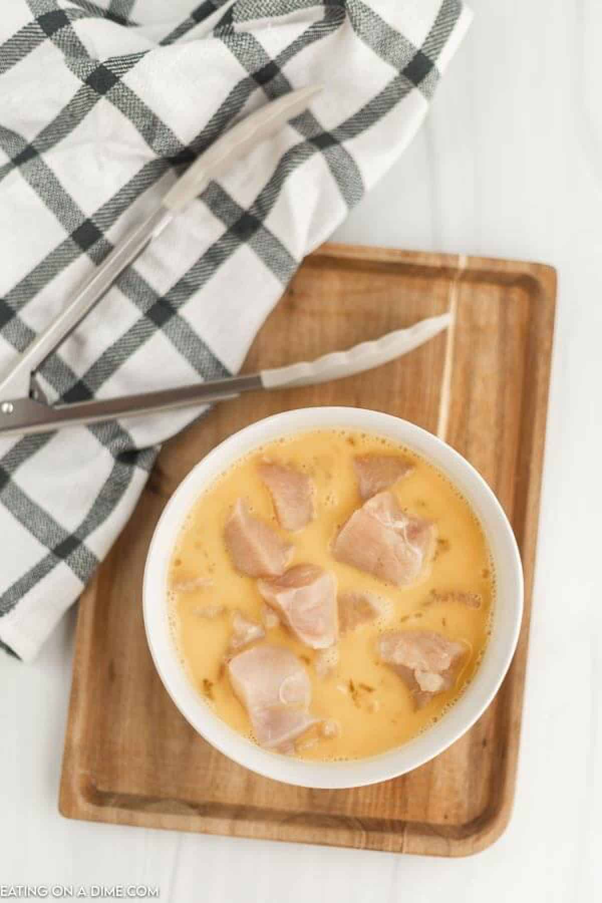 Chicken pieces in a bowl with egg mixture on a cutting board with kitchen tongs