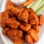 A plate of buffalo chicken bites coated in orange sauce, reminiscent of Air Fryer Boneless Chicken Wings, served with celery, carrot sticks, and a cup of white dipping sauce.