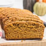 Sliced loaf of Gluten Free Pumpkin Bread on a wooden cutting board, showing a moist, orange-brown interior with a soft texture. Some fall decorations appear blurred in the background.