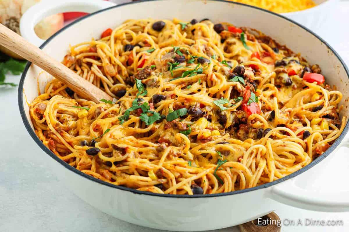 A white pot filled with Mexican Spaghetti, featuring ground beef, black beans, diced tomatoes, corn, and chopped red bell peppers. All topped with melted cheese and fresh parsley. A wooden spoon is placed in the pot for serving this delicious spaghetti dish.