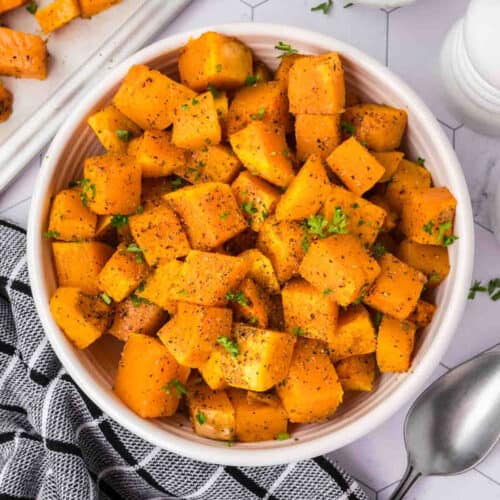 A bowl of roasted butternut squash cubes, baked to perfection and sprinkled with herbs and black pepper. The dish is in a white bowl placed on a light-colored surface. A portion of a black and white patterned cloth and a silver spoon are visible on the side.