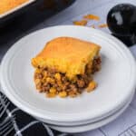 A serving of Cornbread Casserole with a golden topping sits on a white plate, layered with ground beef, corn, and beans.