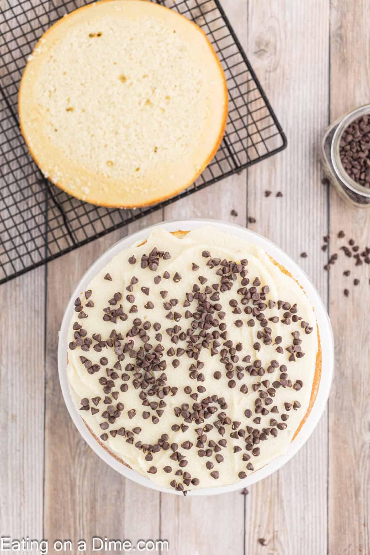 One round cake on a cooling rack with a round cake frosted topped with chocolate chips