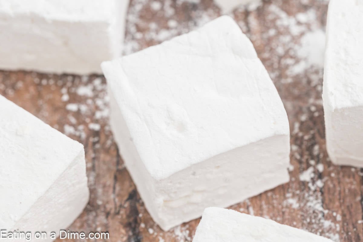 Close-up of DIY marshmallows on a wooden surface, lightly dusted with powdered sugar. These homemade treats boast a soft, fluffy texture and are neatly cut into square shapes.