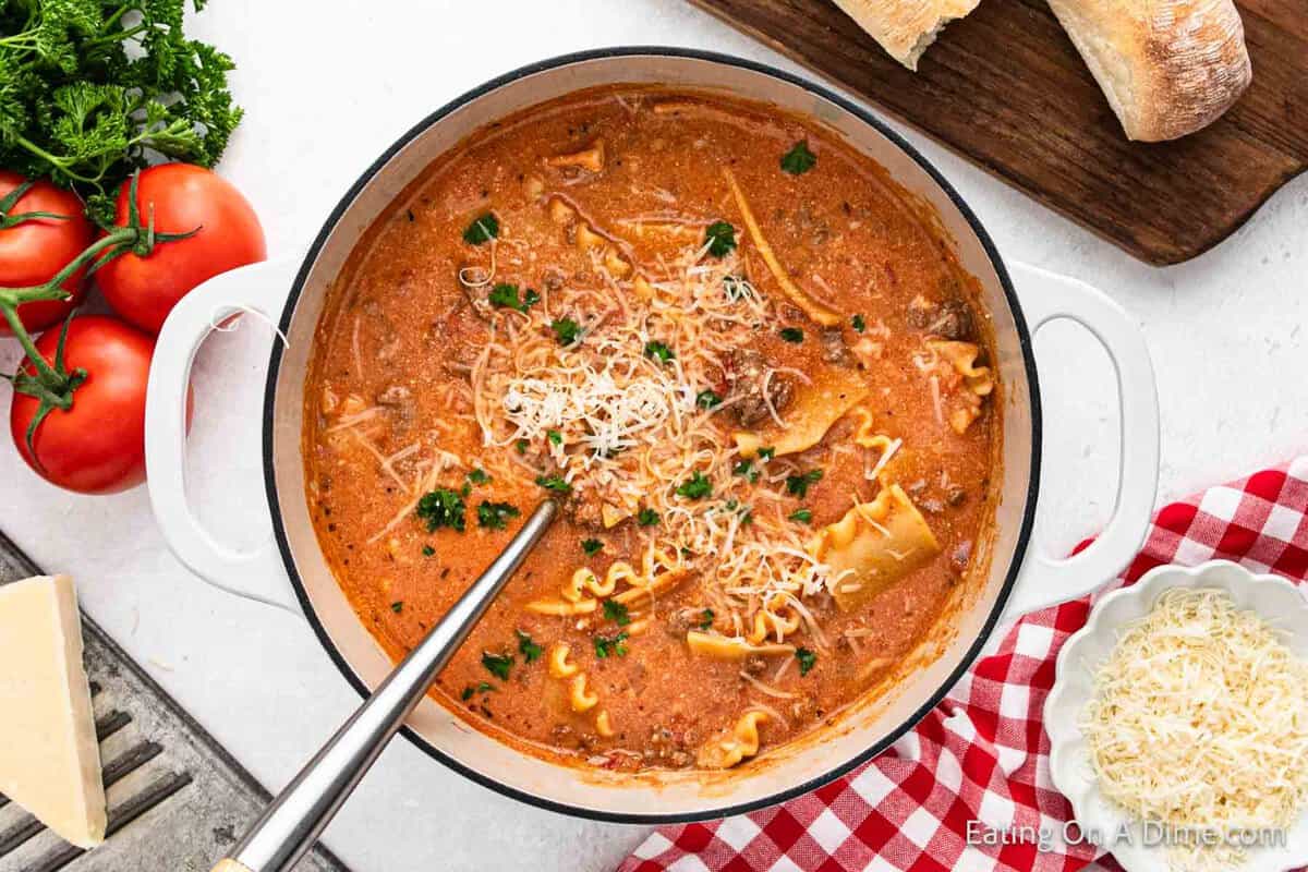 A pot of rich lasagna soup, with pieces of pasta peeking through the creamy broth, is topped with shredded cheese and parsley. Fresh tomatoes, parsley, and slices of bread surround the scene on a checkered cloth.