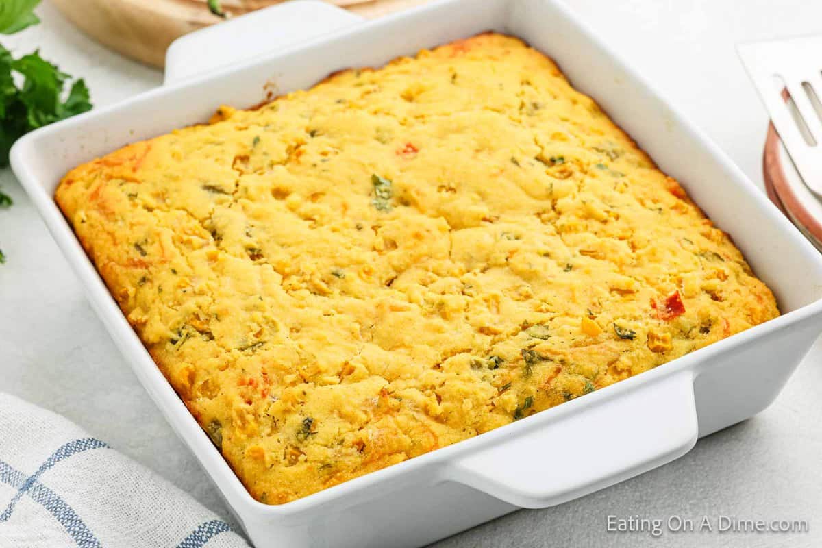 A square dish holds a freshly baked Mexican Cornbread Casserole, adorned with visible pieces of vegetables. The cornbread boasts a golden brown top and sits on a white table beside a blue and white striped cloth.