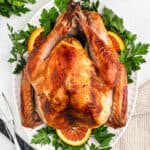 A whole roasted turkey on a white platter, garnished with fresh parsley and orange slices, with serving utensils placed nearby.