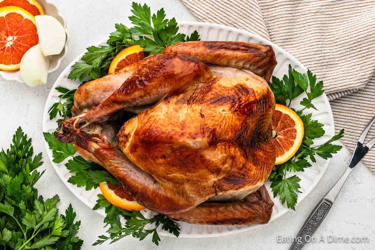 Golden brown whole turkey on a white platter with fresh herbs and slice oranges on the side.