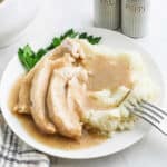A plate of mashed potatoes topped with slices of turkey and rich Turkey Neck Gravy, garnished with fresh parsley, sits next to salt and pepper shakers. A fork rests in the mashed potatoes.