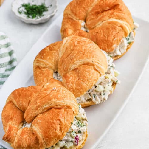 Three croissant sandwiches filled with the best chicken salad recipe is served on a white plate. A small sprig of fresh dill is visible in the background.
