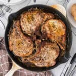 Four seasoned Baked Pork Chops simmer in a cast iron skillet with savory sauce. The skillet rests on a white wooden surface beside a brown checked towel, forks, and a plate of food.