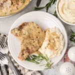 A white plate with Smothered Pork Chops in creamy gravy, served alongside mashed potatoes topped with gravy. Fresh rosemary garnish is on the plate. Utensils, extra mashed potatoes, and salt and pepper shakers are nearby.