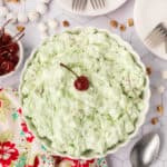 watergate salad topped with a red cherry served in a white bowl. a spoon, some forks over white plates, bowl of cherries, and a flower cloth blurred in the background.