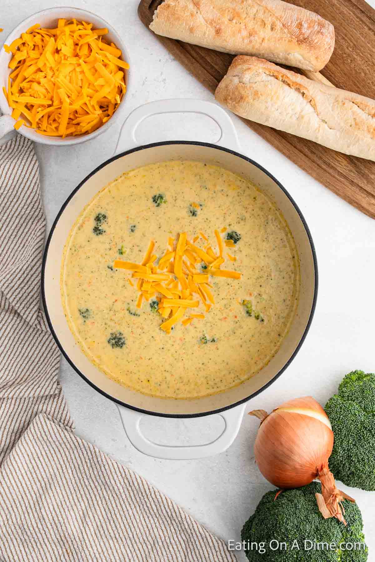 A creamy broccoli cheese soup simmers in a pot, its surface adorned with shredded cheddar. Nearby, a bowl of extra cheese sits beside an onion, a fresh broccoli head, and two crusty baguettes on a wooden board. A striped cloth napkin completes the cozy setting.